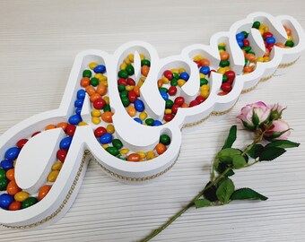 Pode incluir: Um letreiro de madeira branco em forma da palavra "Mãe" cheio de doces coloridos. O letreiro tem uma moldura dourada.
