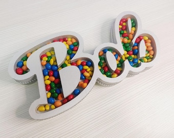 Pode incluir: Uma placa decorativa de madeira cinza que soletra a palavra "Bolo" em uma fonte lúdica. As letras são preenchidas com doces coloridos, incluindo vermelho, amarelo, azul e verde. A placa está em uma superfície branca.
