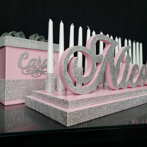 May include: A pink and silver decorative centerpiece with the name "Nicole" in silver glitter. The centerpiece includes white candles and a pink box with the name "Casey" in silver glitter. The base and box are adorned with silver rhinestones.