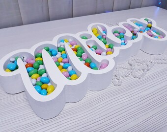 Pode incluir: Tabuleiro de letras de espuma branca que soletra "Mila" cheio de doces em cores pastel. O tabuleiro está sobre uma superfície de madeira branca.