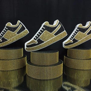 Puede incluir: Tres zapatillas deportivas doradas y negras brillantes con detalles blancos se exhiben sobre tres plataformas texturizadas doradas en niveles. Las zapatillas están ligeramente anguladas, creando un efecto visual dinámico sobre un fondo negro.