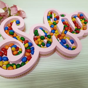 May include: Pink wooden name sign with the name "Sofia" spelled out in a decorative font. The sign is filled with colourful sweets.