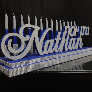 Può includere: Un candelabro di Hanukkah blu e argento con il nome "Nathan" in argento glitterato. Il candelabro ha candele argentate ed è decorato con strass argentati. Lo sfondo è nero.