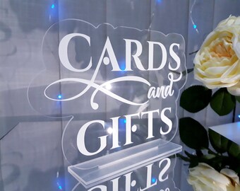 May include: A clear acrylic sign with white lettering that reads "Cards and Gifts". The sign is illuminated with blue lights and is sitting on a white stand.