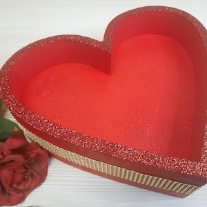 Op de afbeelding: Rode hartvormige doos met een gouden glitterrand. De doos is gemaakt van karton en heeft een rode glitterafwerking.