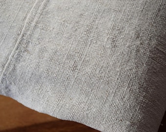 May include: Close-up of a light gray, textured fabric. The material has a woven appearance with visible threads and a subtle, natural color variation. The fabric is folded, showing a seam running vertically.