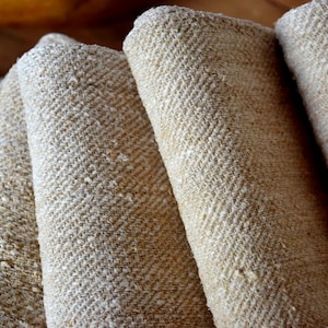 Könnte beinhalten: Nahaufnahme von mehreren Rollen beigefarbenem, strukturiertem Stoff. Der Stoff hat ein gewebtes Fischgrätenmuster. Die Rollen sind eng gewickelt und zeigen die Kanten des Stoffes. Die Beleuchtung hebt die Textur und Farbvariationen hervor.