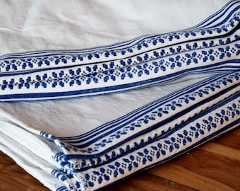 May include: White linen fabric with blue striped and floral patterned trim. The fabric is folded, showcasing the detailed design and texture. The blue and white color scheme creates a classic, clean aesthetic.