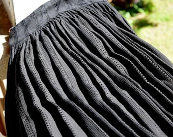 May include: Close-up of a black pleated skirt. The fabric has a textured, ribbed appearance with small, evenly spaced perforations. The skirt's waistband is visible at the top, and the pleats cascade downwards, creating a flowing effect. The skirt is made of a lightweight material.
