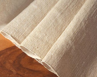 May include: Close-up of folded, off-white fabric with a textured weave. The fabric is arranged in soft folds, revealing the raw edges. The neutral color and texture suggest a natural fiber textile, possibly linen or cotton.