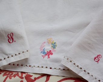 May include: Two white linen tea towels with embroidered details. One towel has a wreath of flowers and the other has the letter "S" embroidered in pink thread.