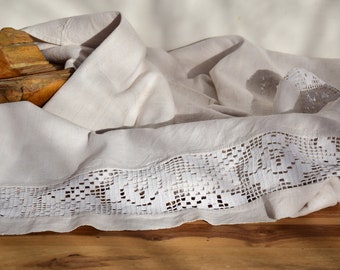 May include: A white linen tablecloth with a delicate lace trim. The tablecloth is draped over a wooden surface.