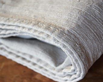 May include: Close-up of a folded, light gray linen textile. The fabric has a textured weave with visible threads and subtle variations in color. The edges are neatly finished with delicate stitching, suggesting a handcrafted item.