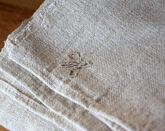 May include: Close-up of a stack of beige linen napkins with a rough, textured weave. The napkins have a simple, rustic aesthetic, with a small, embroidered design in the corner. The edges are frayed, adding to the handmade feel.