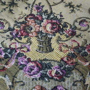 May include: Close-up of a tapestry with a floral design. The tapestry features a central arrangement of flowers in a basket, with pink and purple roses. The background is a light beige colour with decorative scrollwork.