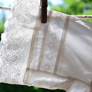 以下が含まれることがあります： 繊細なレースのトリムと刺繍された花の模様が施された白い綿の生地。生地は物干しロープにかけられています。