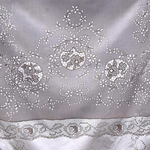 May include: A gray textile with white floral embroidery and a lace border. The design features three central floral medallions surrounded by delicate white floral and leaf patterns. The lace border adds a vintage touch to the fabric.