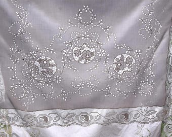 May include: A grey textile with white floral embroidery and a lace border. The design features three central floral medallions surrounded by delicate white floral and leaf patterns. The lace border adds a vintage touch to the fabric.