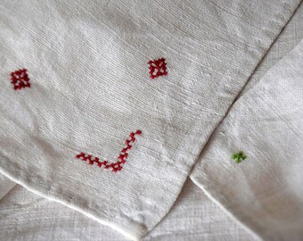 May include: Close-up of a white linen fabric with red and green cross-stitch embroidery. The embroidery features small square and angular designs. The fabric is folded, showing texture and detail.
