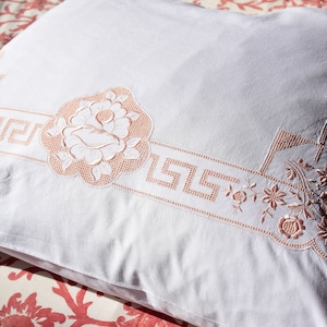 May include: A white pillowcase with a delicate pink embroidered design featuring a floral motif and a Greek key pattern.