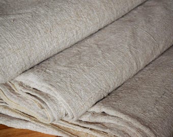 May include: A folded length of heavy-weight, natural-colored linen fabric with a visible, coarse weave. The material displays an undyed, beige-toned appearance with subtle variations in the fiber texture, showcasing a dense and durable textile construction.