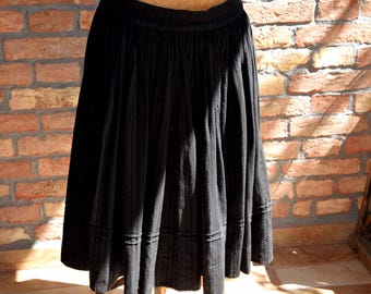May include: A black skirt with a textured, pleated design. The skirt has a wide waistband and a flared silhouette, with a decorative band near the hem. The skirt is displayed against a brick wall.