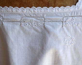 Puede incluir: Primer plano de una tela de lino blanca con intrincados bordados. El borde superior presenta un diseño festoneado. El bordado incluye patrones florales y geométricos, con un motivo circular central. La tela parece ser parte de un textil vintage.