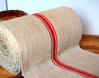 May include: A roll of natural-colored burlap fabric with a bold red stripe down the center. The fabric is tightly woven and appears to be new. The roll is resting on a wooden surface.