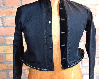 May include: A black long-sleeve jacket featuring a subtle dotted pattern, a high collar, and a front button closure. The garment includes a decorative pleated trim along the bottom hem and vertical textured detailing along the button placket.