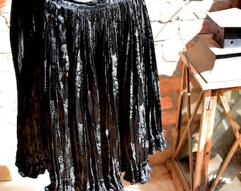 May include: A black, flowing skirt with a floral pattern. The fabric has a shiny, textured appearance, with vertical pleats adding to the design. The skirt has a ruffled hemline and is hanging in front of a brick wall.