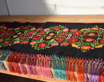 May include: Black table runner with colorful embroidered floral designs and a fringe border. The runner features red, green, yellow, blue, and purple flowers.