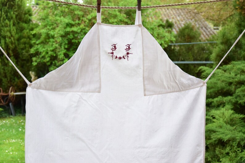 May include: A white linen apron with red embroidered floral details and the letters "JJ" in the center.