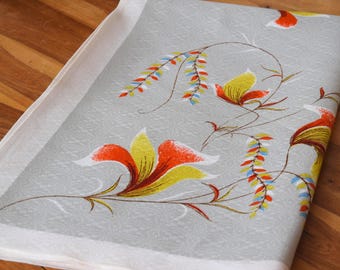 May include: A folded, light gray fabric with a floral pattern. The design features orange, yellow, and white flowers with brown stems and accents. The fabric has a white border and is set on a wooden surface.