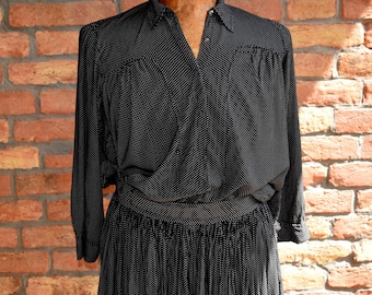 May include: Black vintage dress with white polka dots. The dress features a collared neckline, long sleeves, and a pleated skirt. The dress is displayed against a brick wall background.
