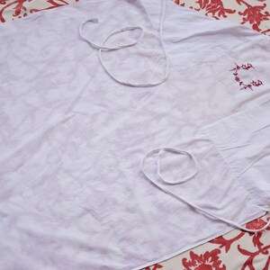 May include: A white apron with a red embroidered design. The apron has two white ties.