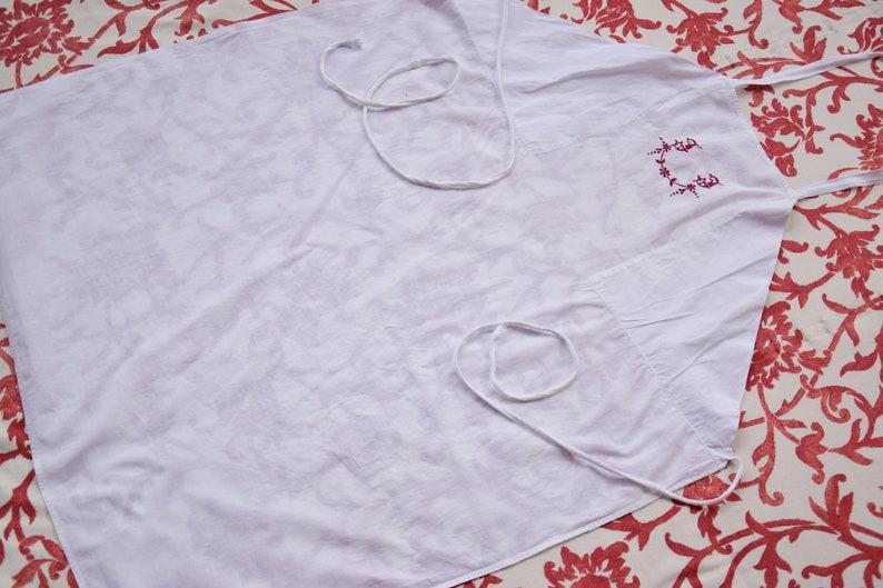 May include: A white apron with a red embroidered design. The apron has two white ties.