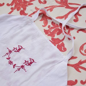 May include: White cotton apron with a pink embroidered floral design. The apron has a single strap that goes over the head.