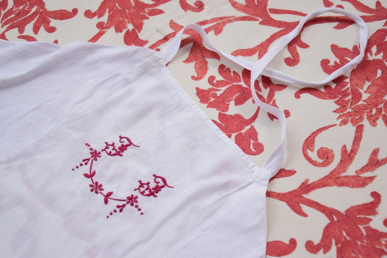 May include: White cotton apron with a pink embroidered floral design. The apron has a single strap that goes over the head.