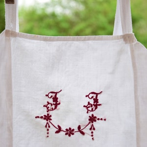 May include: White linen apron with red embroidered initials "JJ" and a floral design.