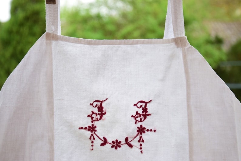 May include: White linen apron with red embroidered initials "JJ" and a floral design.