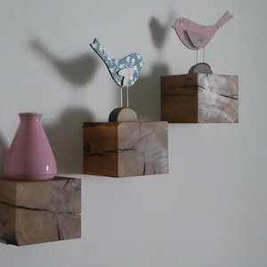 Op de afbeelding: Drie houten planken met een roze vaas op de onderste plank en twee houten vogels op de bovenste twee planken. De planken zijn gemaakt van rustiek hout met een natuurlijke afwerking.