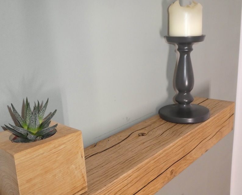 Puede incluir: Una estanter&iacute;a de madera con un acabado de madera natural y una peque&ntilde;a planta suculenta en una maceta de madera. Un candelero negro con una vela blanca est&aacute; en la estanter&iacute;a.