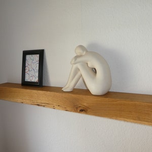 Puede incluir: Una estanter&iacute;a de madera con una escultura de cer&aacute;mica blanca de una persona sentada sobre ella. Un cuadro enmarcado con un dise&ntilde;o abstracto de colores est&aacute; en el lado izquierdo de la estanter&iacute;a.