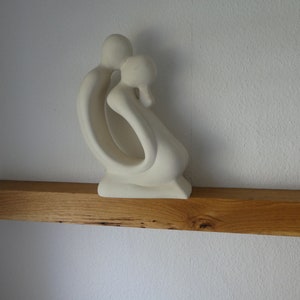Puede incluir: Escultura de cer&aacute;mica blanca de dos figuras abraz&aacute;ndose, expuesta en una estanter&iacute;a de madera.