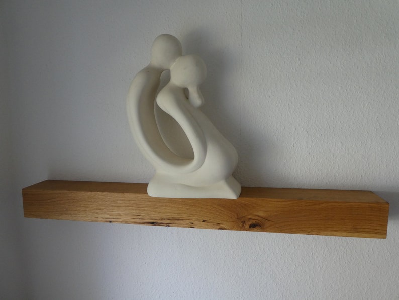 Puede incluir: Escultura de cer&aacute;mica blanca de dos figuras abraz&aacute;ndose, expuesta en una estanter&iacute;a de madera.