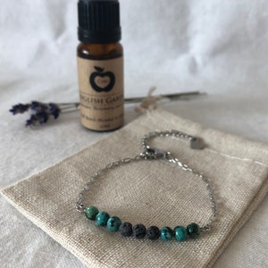 May include: A silver chain bracelet with turquoise and black lava rock beads. The bracelet is on a beige fabric surface. A small bottle of essential oil is in the background.
