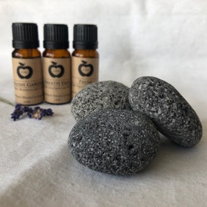 May include: Three amber glass essential oil bottles with black lids, labelled "English Garden", "Breath Easy", and "Pause". Beside them are four grey, textured stones and a sprig of lavender. The items are arranged on a white linen surface.