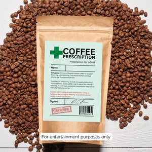 Könnte beinhalten: Ein brauner Papierbeutel mit einem grün-weißen Etikett, auf dem "Kaffee-Rezept" steht, mit der Rezeptnummer A2468. Das Etikett enthält auch eine tägliche Dosis, mögliche Nebenwirkungen und einen Haftungsausschluss. Der Beutel ist von Kaffeebohnen umgeben.
