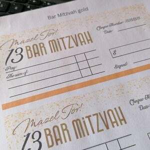 May include: A white paper sheet featuring two printed Bar Mitzvah gift check templates with gold-colored speckle accents. The text includes "Mazel Tov!", "13 BAR MITZVAH", "Pay:", "The sum of:", "Cheque Number:", "Date:", "$", and "Signed:".