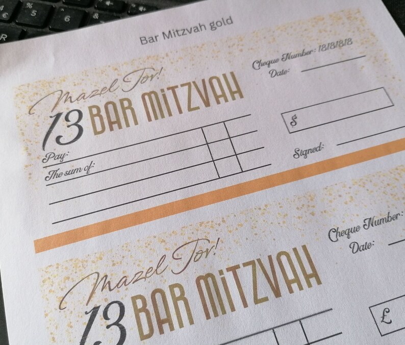 May include: A white paper sheet featuring two printed Bar Mitzvah gift check templates with gold-colored speckle accents. The text includes "Mazel Tov!", "13 BAR MITZVAH", "Pay:", "The sum of:", "Cheque Number:", "Date:", "$", and "Signed:".
